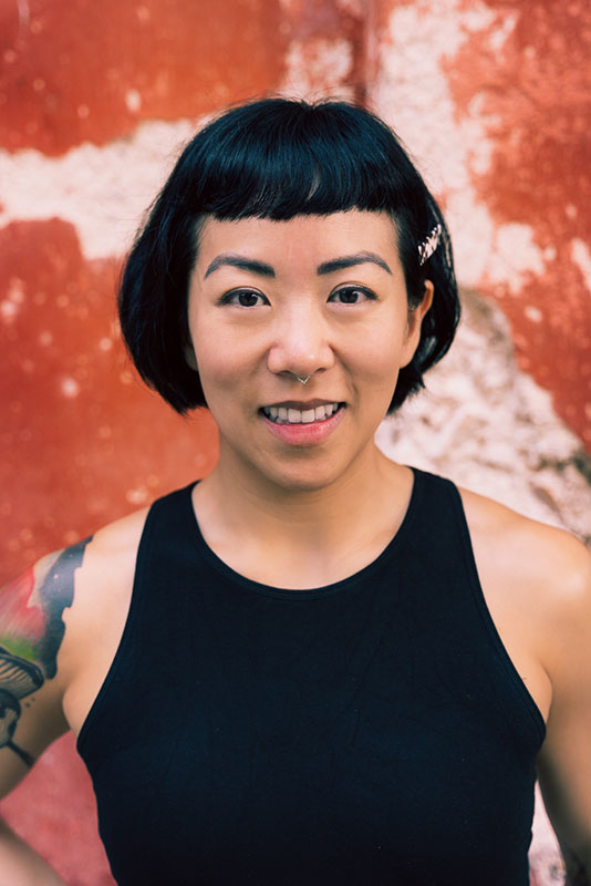 Melody Li's Headshot: middle-age East Asian femme-presenting human with medium tone skin, bob haircut, wearing a black tank top, tattoo on right shoulder, smiling at the viewer.